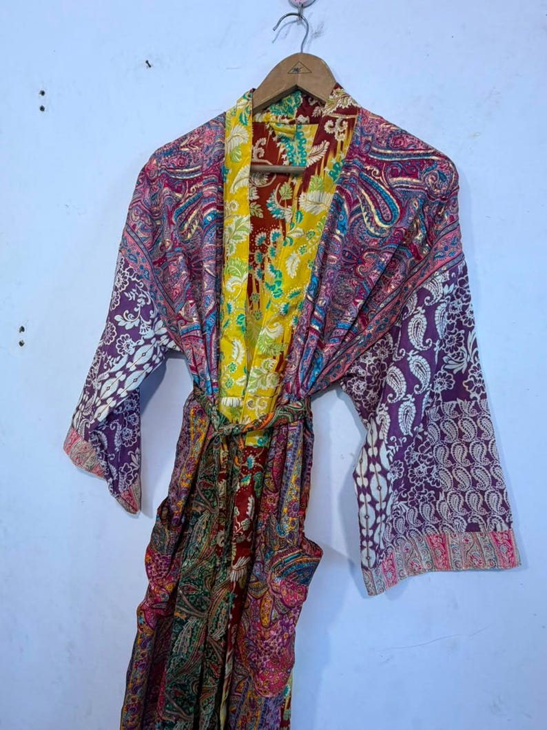 Może przedstawiać: Szlafrok typu kimono z długimi rękawami, ozdobiony żywym wzorem patchworkowym z r&oacute;żnymi motywami kwiatowymi i paisley w odcieniach ż&oacute;łci, głębokiej czerwieni, fioletu i r&oacute;żu. Odzież posiada pasujący pasek z tkaniny oraz jaskrawoż&oacute;łty kołnierz wewnętrzny.
