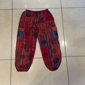May include: Red and multi-colored patchwork pants featuring a drawstring waist, elasticized cuffs, and two side cargo pockets. The fabric displays a variety of vertical striped patterns and black circular graphic prints throughout the garment.