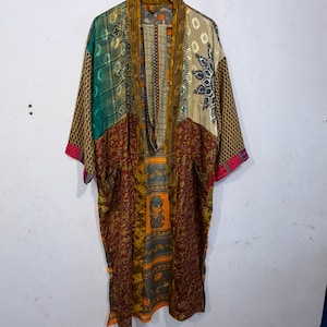 May include: A patchwork kimono robe with a variety of patterns and colors, including green, gold, red, and beige. The robe is hanging on a wooden hanger against a white wall. The sleeves have a pink trim.
