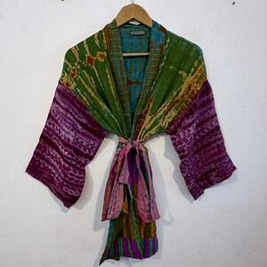 May include: A colorful, tie-dye kimono robe with a belt. The robe features shades of green, purple, and blue. The sleeves are a deep purple, and the belt is a mix of pink, green, and purple. The robe is hanging on a wooden hanger.