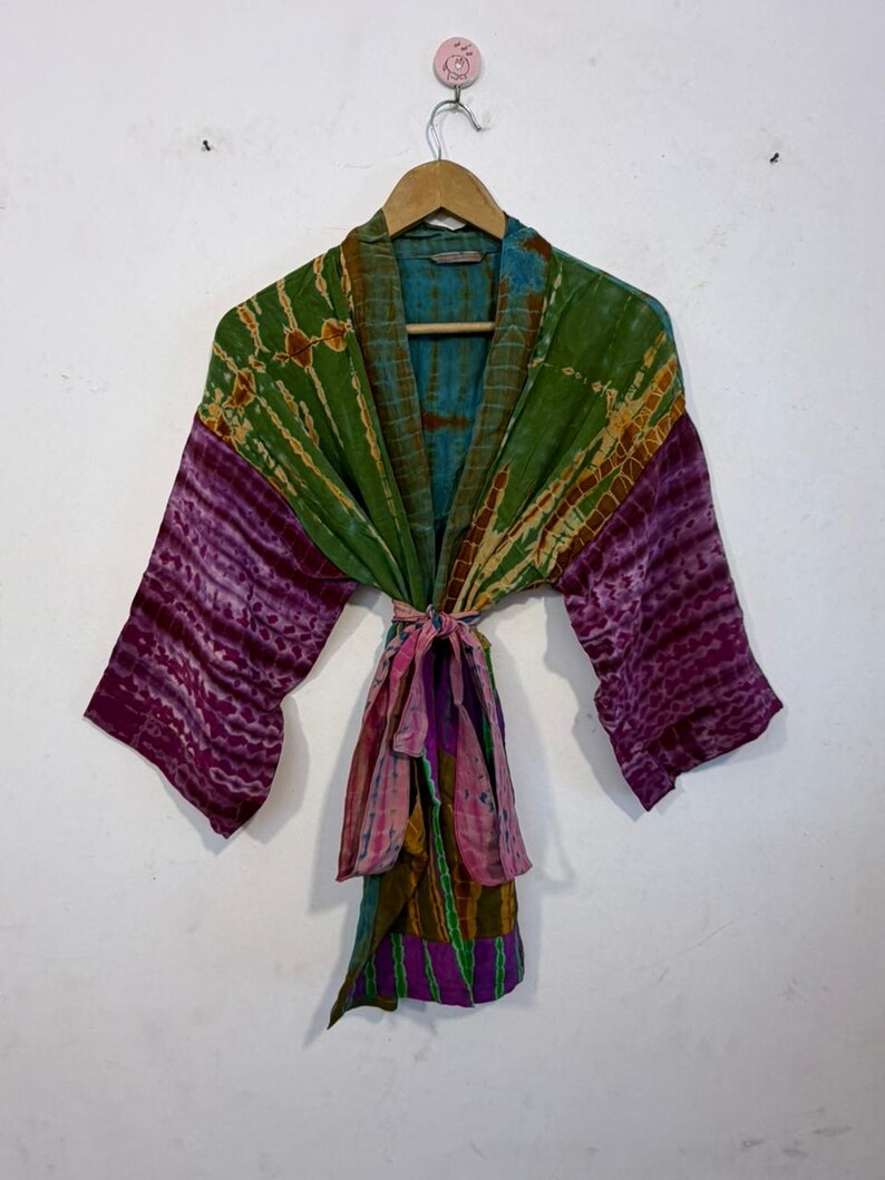 May include: A colorful, tie-dye kimono robe with a belt. The robe features shades of green, purple, and blue. The sleeves are a deep purple, and the belt is a mix of pink, green, and purple. The robe is hanging on a wooden hanger.