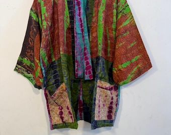 May include: A kimono-style jacket featuring a patchwork design with various tie-dye patterns in shades of green, rust orange, purple, and blue. The garment has wide sleeves, an open front, and two rectangular patch pockets on the lower front.