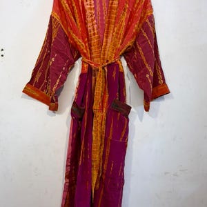 May include: A long-sleeved robe featuring a vibrant tie-dye pattern in shades of magenta, orange, and yellow. The garment includes a matching fabric waist tie, an open front design, and two front pockets, all displayed on a wooden hanger.