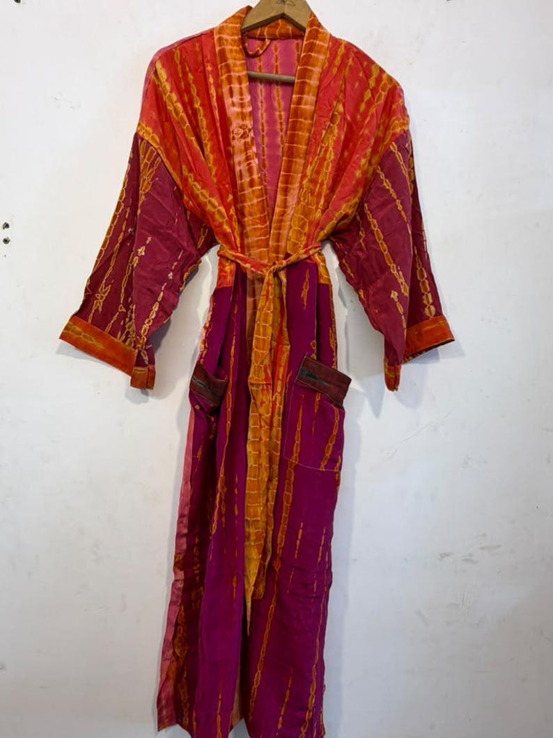 May include: A long-sleeved robe featuring a vibrant tie-dye pattern in shades of magenta, orange, and yellow. The garment includes a matching fabric waist tie, an open front design, and two front pockets, all displayed on a wooden hanger.