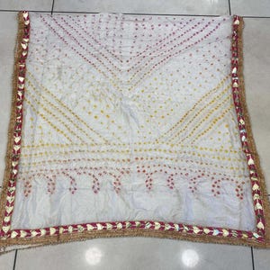 May include: White scarf with a patterned design in yellow and red dots. The scarf has a decorative border with gold and red accents. The scarf is made of a soft, flowing fabric.