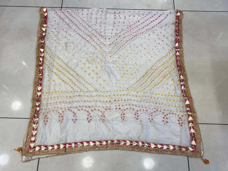 May include: White scarf with a patterned design in yellow and red dots. The scarf has a decorative border with gold and red accents. The scarf is made of a soft, flowing fabric.