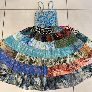 Puede incluir: Un vestido patchwork con un corpi&ntilde;o azul con estampado floral y tirantes finos. La falda presenta un dise&ntilde;o escalonado con varias telas estampadas en tonos azules, verdes y naranjas. El vestido est&aacute; tendido sobre una superficie embaldosada.