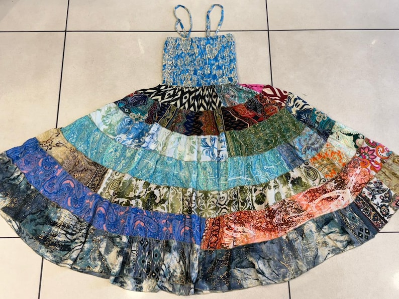 Puede incluir: Un vestido patchwork con un corpi&ntilde;o azul con estampado floral y tirantes finos. La falda presenta un dise&ntilde;o escalonado con varias telas estampadas en tonos azules, verdes y naranjas. El vestido est&aacute; tendido sobre una superficie embaldosada.