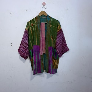 May include: A colorful, patterned kimono-style robe with a tie-dye effect. The robe features shades of green, purple, and pink, with pockets and a matching sash. It is hanging on a wooden hanger against a white wall.