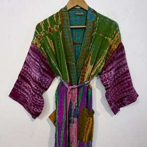 May include: A colorful tie-dye robe with a belt, hanging on a wooden hanger. The robe features shades of green, purple, and blue. The sleeves are a deep purple, and the body has a mix of colors. It has two pockets.