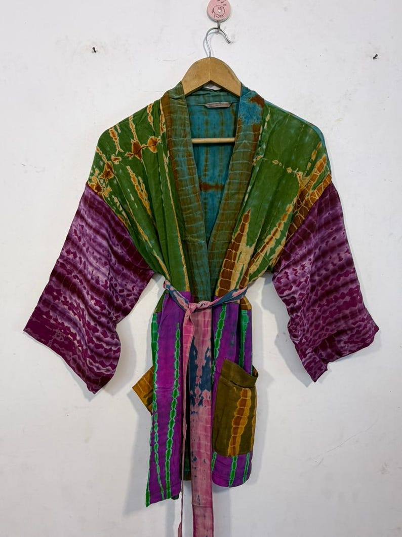 May include: A colorful tie-dye robe with a belt, hanging on a wooden hanger. The robe features shades of green, purple, and blue. The sleeves are a deep purple, and the body has a mix of colors. It has two pockets.