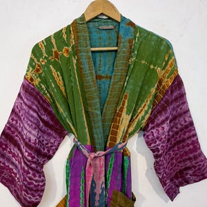 May include: A tie-dye robe in shades of green, purple, and blue hangs on a wooden hanger. The robe features long sleeves with a purple pattern, a green bodice, and a colorful belt. The robe has pockets.