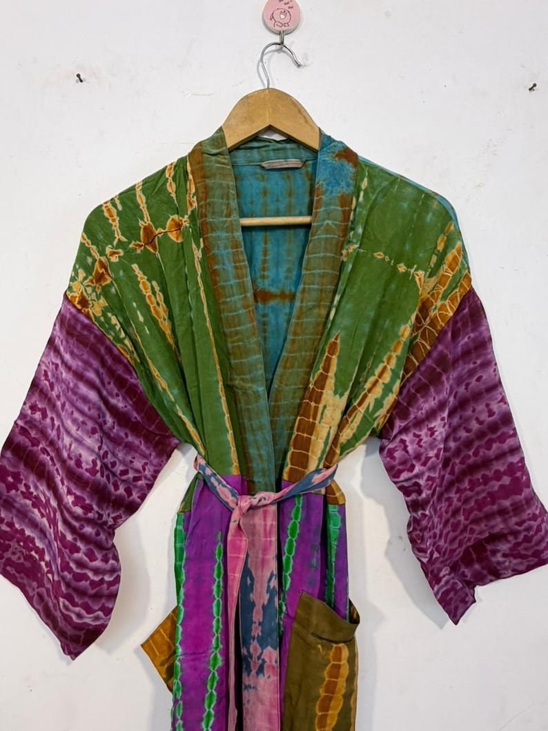May include: A tie-dye robe in shades of green, purple, and blue hangs on a wooden hanger. The robe features long sleeves with a purple pattern, a green bodice, and a colorful belt. The robe has pockets.