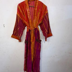 May include: A long-sleeved robe featuring a vibrant tie-dye pattern in shades of magenta and orange. The garment includes a matching tie belt at the waist, a wide collar, and two front pockets, all hanging from a wooden hanger.