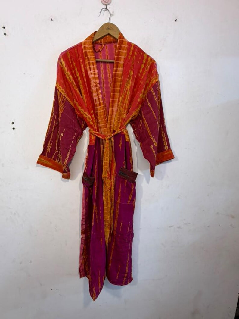 May include: A long-sleeved robe featuring a vibrant tie-dye pattern in shades of magenta and orange. The garment includes a matching tie belt at the waist, a wide collar, and two front pockets, all hanging from a wooden hanger.