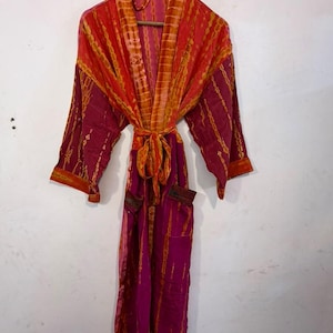 May include: A long-sleeved robe featuring a vibrant tie-dye pattern in shades of magenta and orange. The garment includes a matching tie belt at the waist, a shawl collar, and two front pockets, all hanging from a wooden hanger.