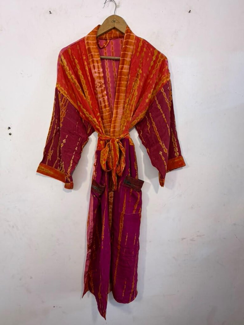 May include: A long-sleeved robe featuring a vibrant tie-dye pattern in shades of magenta and orange. The garment includes a matching tie belt at the waist, a shawl collar, and two front pockets, all hanging from a wooden hanger.