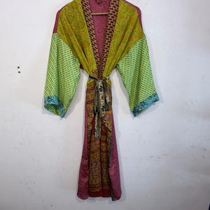 May include: A colorful, patterned robe with a tie belt, hanging on a wooden hanger. The robe features a mix of green, yellow, pink, and brown fabrics. The sleeves are a lighter green with a blue trim. The robe is made of a lightweight material.