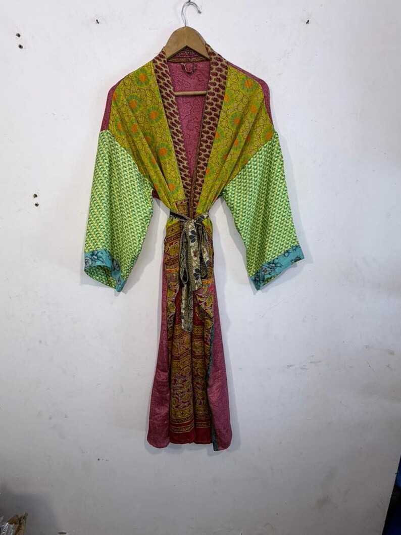 May include: A colorful, patterned robe with a tie belt, hanging on a wooden hanger. The robe features a mix of green, yellow, pink, and brown fabrics. The sleeves are a lighter green with a blue trim. The robe is made of a lightweight material.