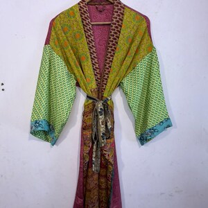May include: A colorful, patterned robe with long sleeves, hanging on a wooden hanger. The robe features a mix of green, yellow, and pink fabrics with various designs. A patterned belt cinches the waist. The sleeves have a green and blue patterned design.