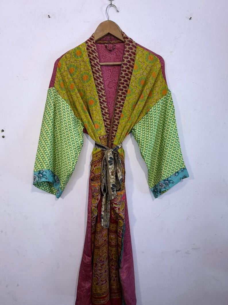 May include: A colorful, patterned robe with long sleeves, hanging on a wooden hanger. The robe features a mix of green, yellow, and pink fabrics with various designs. A patterned belt cinches the waist. The sleeves have a green and blue patterned design.