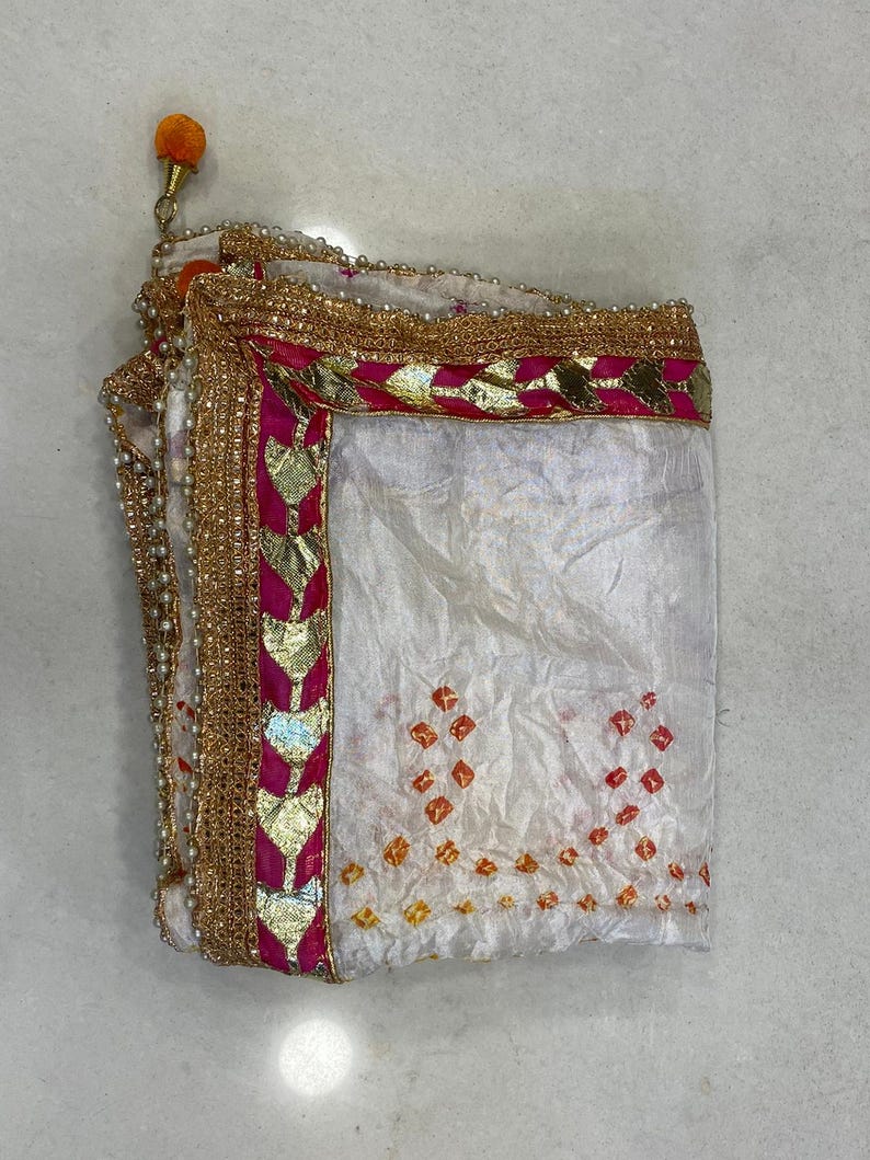 May include: White fabric with a decorative border featuring gold, pink, and pearl accents. The fabric has a pattern of orange and red diamond shapes. An orange tassel is attached to the corner.