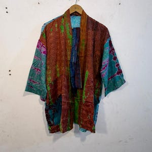May include: A kimono-style jacket featuring a patchwork design with various patterns and colors including rust orange, teal, green, and purple. The garment has an open front, wide three-quarter length sleeves, and a lightweight, textured fabric construction.