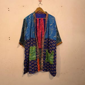Könnte beinhalten: Eine farbenfrohe Kimono-Jacke mit Muster und kurzen Ärmeln. Die Jacke besteht aus einer Mischung aus blauen, grünen und rosa Stoffen mit Blumen- und Paisley-Mustern. Sie hat zwei Vordertaschen und hängt an einem Holzbügel.