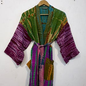 May include: A colorful, tie-dye patterned robe with long sleeves and a matching belt. The robe features shades of green, purple, and blue. It has two pockets and is hanging on a wooden hanger against a white wall.