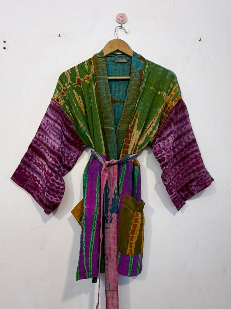 May include: A colorful, tie-dye patterned robe with long sleeves and a matching belt. The robe features shades of green, purple, and blue. It has two pockets and is hanging on a wooden hanger against a white wall.