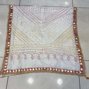 May include: White scarf with a patterned design in yellow and red dots. The scarf has a decorative border with gold and red accents. The scarf is made of a lightweight fabric.