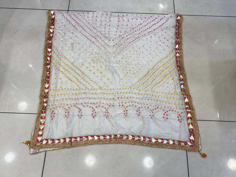 May include: White scarf with a patterned design in yellow and red dots. The scarf has a decorative border with gold and red accents. The scarf is made of a lightweight fabric.