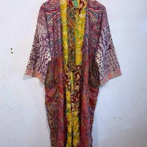 Może przedstawiać: Szlafrok w stylu kimono z długimi rękawami, ozdobiony żywym wzorem patchworkowym z r&oacute;żnymi motywami kwiatowymi i paisley w odcieniach ż&oacute;łci, głębokiej czerwieni, fioletu i r&oacute;żu. Odzież ma otwarty prz&oacute;d i jest zawieszona na drewnianym wieszaku.