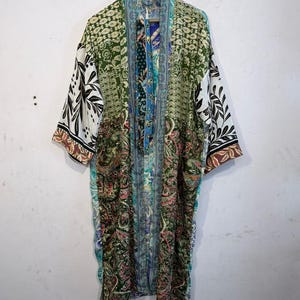 Può includere: Un accappatoio kimono a maniche lunghe caratterizzato da un design patchwork con vari motivi floreali e paisley nei toni del verde, blu e bianco. Il capo presenta un'apertura frontale, un colletto ampio e maniche larghe con bordi.