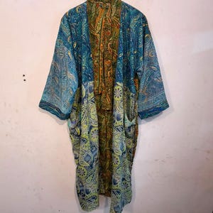May include: A long-sleeved kimono-style robe featuring a patchwork design of blue, green, and gold paisley and floral patterns. The garment has an open front, wide sleeves, and two visible front pockets, hanging on a wooden hanger against a wall.