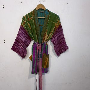 May include: A colorful tie-dye kimono robe with a belt, hanging on a wooden hanger. The robe features shades of green, purple, and blue. The sleeves and belt are a deep purple, while the body has a mix of green and blue.