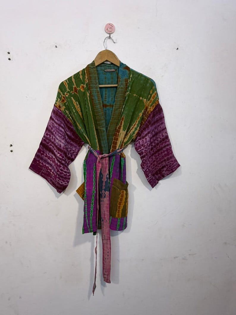 May include: A colorful tie-dye kimono robe with a belt, hanging on a wooden hanger. The robe features shades of green, purple, and blue. The sleeves and belt are a deep purple, while the body has a mix of green and blue.