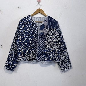 May include: A patchwork quilted jacket in blue and white. The jacket displays a variety of patterns, including floral and geometric designs. It has long sleeves and a V-neck, and is suspended from a wooden hanger.