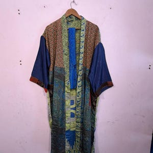 Può includere: Una lunga vestaglia kimono patchwork con una varietà di motivi e colori. La vestaglia presenta un mix di disegni floreali, paisley e geometrici nelle tonalità del blu, verde, marrone e arancione. Le maniche sono blu navy tinta unita. La vestaglia è appesa a una gruccia di legno.
