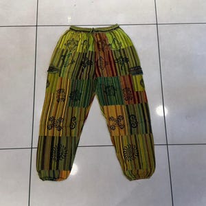 May include: Loose-fitting pants featuring a patchwork design with vertical stripes in shades of lime green, yellow, and brown. The fabric is decorated with black printed symbols, including suns and geometric shapes, and includes an elastic waistband.