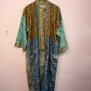 May include: A long-sleeved kimono robe featuring a patchwork design of various paisley and geometric patterns in shades of teal, blue, olive green, and gold. The garment has an open front, wide sleeves, and a matching fabric belt hanging down.