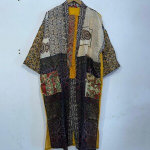 Könnte beinhalten: Kimono-ähnlicher Bademantel im Patchwork-Stil mit gelbem Futter und Besatz. Der Bademantel weist eine Vielzahl von gemusterten Stoffen in Creme-, Schwarz-, Rot- und Gelbtönen auf. Der Bademantel hängt an einem Holzbügel vor einer weißen Wand.