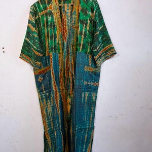 May include: A long-sleeved kimono-style robe featuring a vibrant tie-dye pattern in shades of emerald green, teal, and burnt orange. The garment has an open front, a wide collar, and two patch pockets on the lower front section of the fabric.