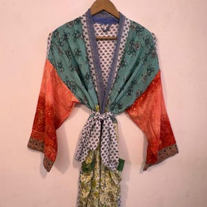 May include: A patterned robe with a tie closure, hanging on a wooden hanger. The robe features panels of teal, orange, and patterned fabric. The sleeves are orange with a red floral design. The tie is white with a blue heart pattern.