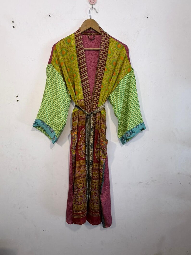 May include: A colorful, patterned kimono robe with a belt. The robe features a mix of colors, including green, yellow, red, and pink. The sleeves have a green pattern, and the body has a floral design. The robe is hanging on a wooden hanger.