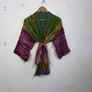 May include: A colorful tie-dye kimono robe with long sleeves. The robe features a mix of green, purple, and pink hues. It is tied at the waist with a matching fabric belt. The robe is hanging on a wooden hanger against a white wall.