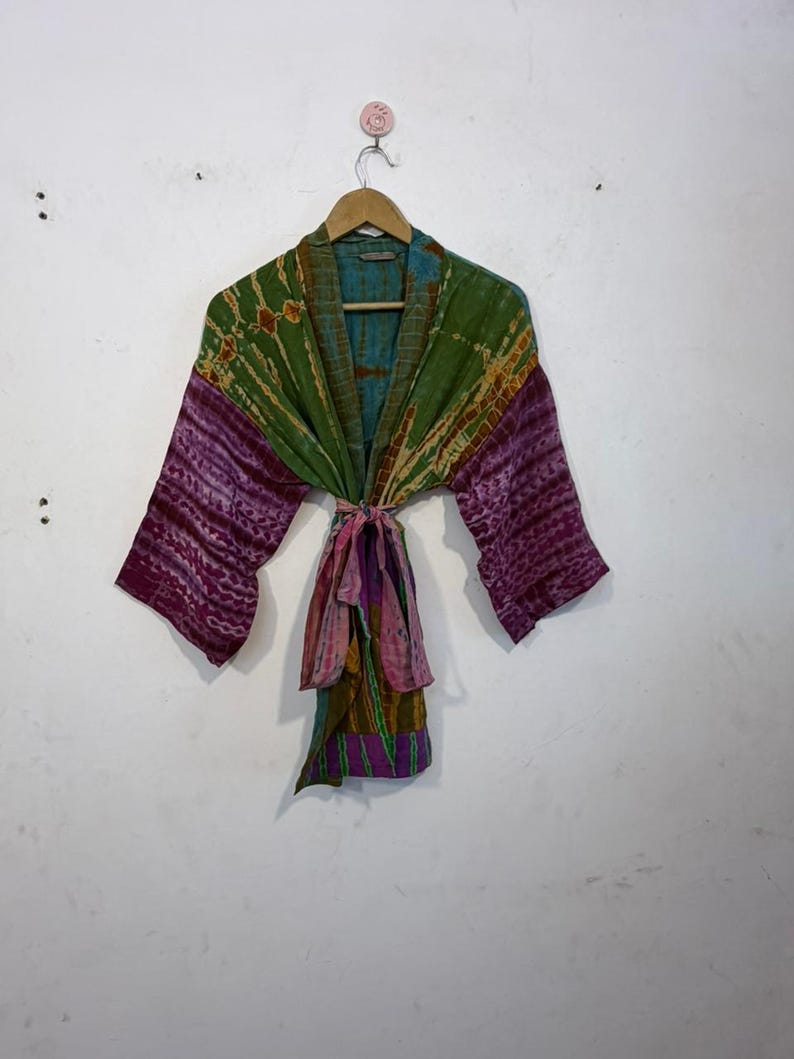 May include: A colorful tie-dye kimono robe with long sleeves. The robe features a mix of green, purple, and pink hues. It is tied at the waist with a matching fabric belt. The robe is hanging on a wooden hanger against a white wall.
