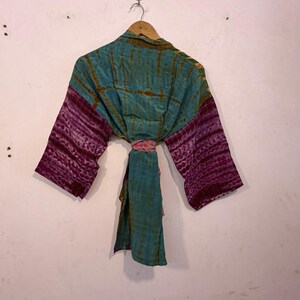 May include: A tie-dye kimono in teal and purple hues hangs on a wooden hanger. The sleeves are a deep purple with a textured pattern, while the body is teal with vertical stripes. A pink sash cinches the waist, adding a pop of color.