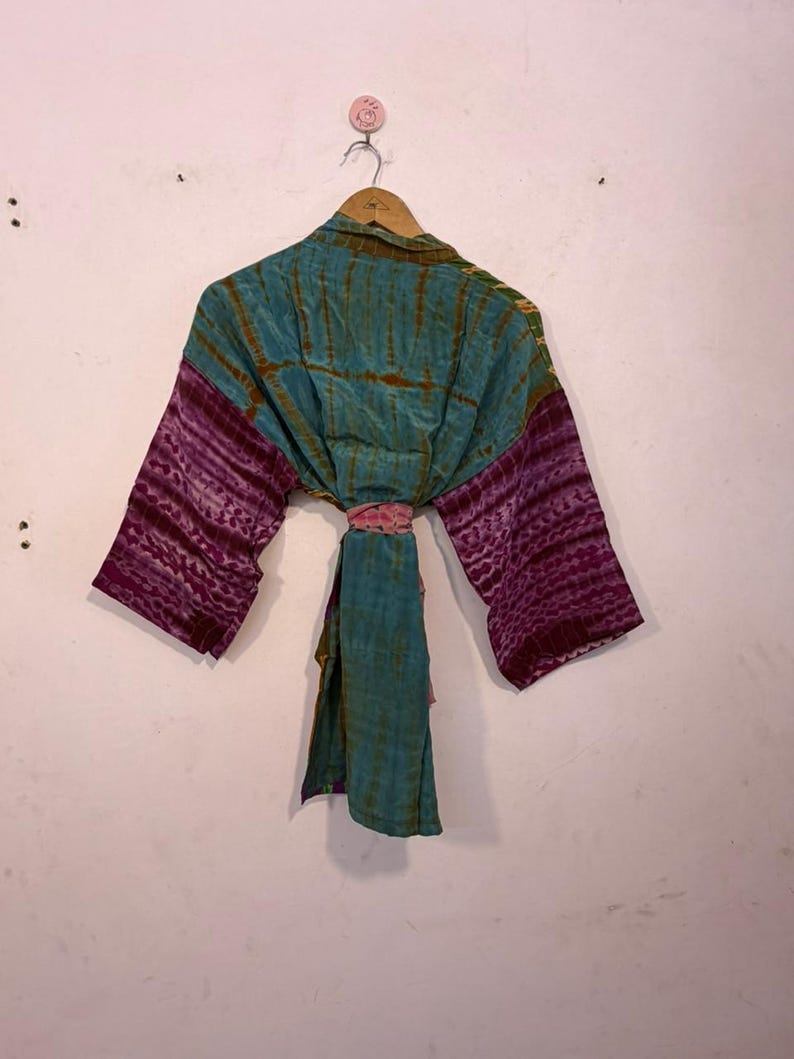 May include: A tie-dye kimono in teal and purple hues hangs on a wooden hanger. The sleeves are a deep purple with a textured pattern, while the body is teal with vertical stripes. A pink sash cinches the waist, adding a pop of color.