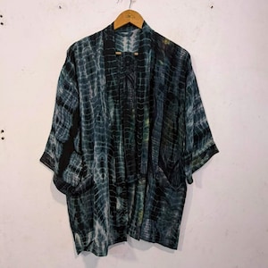 May include: An open-front kimono jacket featuring a dark teal and black tie-dye pattern with vertical stripes. The garment has wide three-quarter length sleeves, a shawl collar, and two front pockets, hanging from a wooden hanger against a wall.